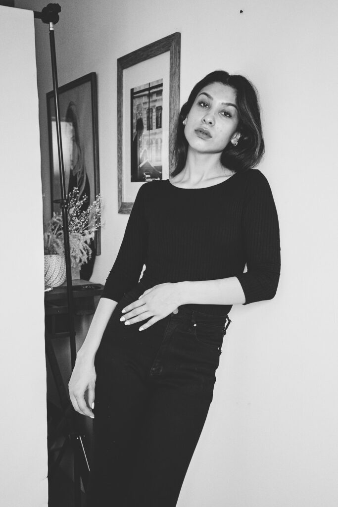 Nadya Inzunza is shown in a monochrome setting leaning against a wall with framed artworks behind her. She wears a black top and black trousers and her pose exudes confidence.